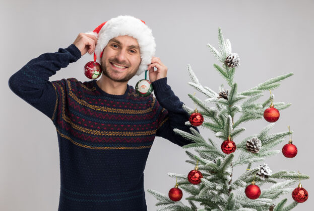 靠近戴着圣诞帽的年轻高加索男子微笑着站在圣诞树旁望着 拿着圣诞装饰球站在隔离在白色墙上的耳朵旁微笑站着舞会