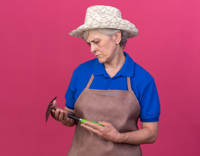 穿自信的上了年纪的女园丁戴着园艺帽拿着锄头看耙子女老人耙