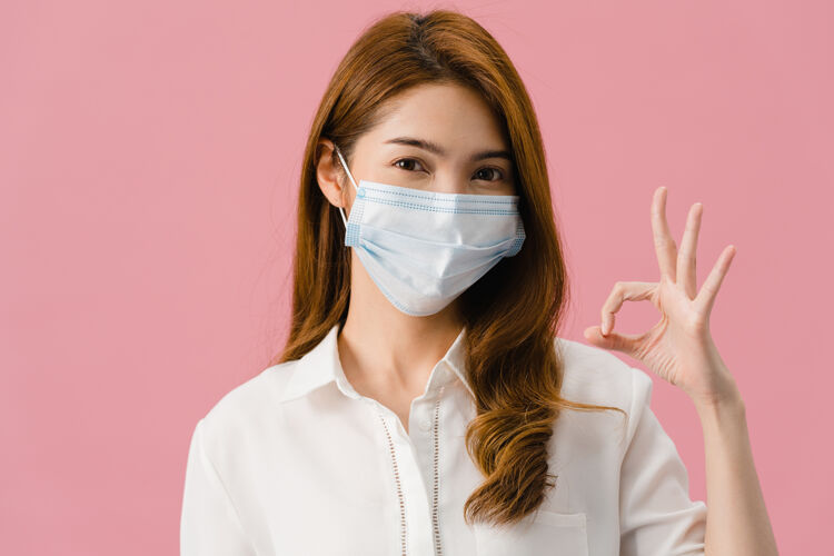 过敏年轻的亚洲女孩戴着医用面罩 穿着休闲服 打着ok的手势 看着隔离在粉色背景上的相机卫生学生医学