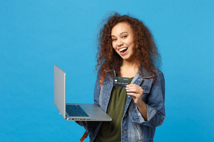 包年轻的非洲裔美国女孩十几岁的学生在牛仔服装 背包工作的电脑 持有银行卡隔离在蓝色的墙壁上支付获奖者青少年