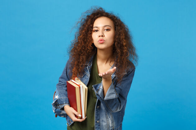 人穿着牛仔服 背着书包的年轻女学生把书孤立地放在蓝色的墙上发送大学班级
