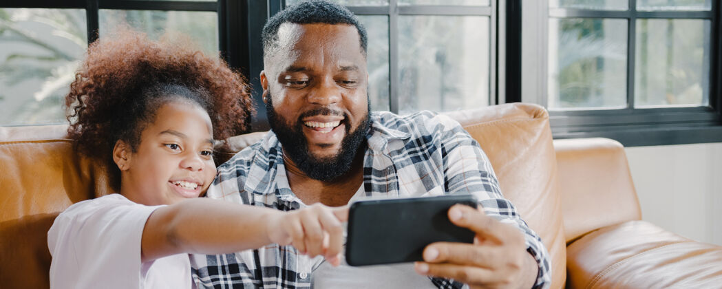 黑人快乐的非洲裔美国家庭爸爸和女儿在家里的沙发上玩得开心 用手机视频通话人休闲客厅