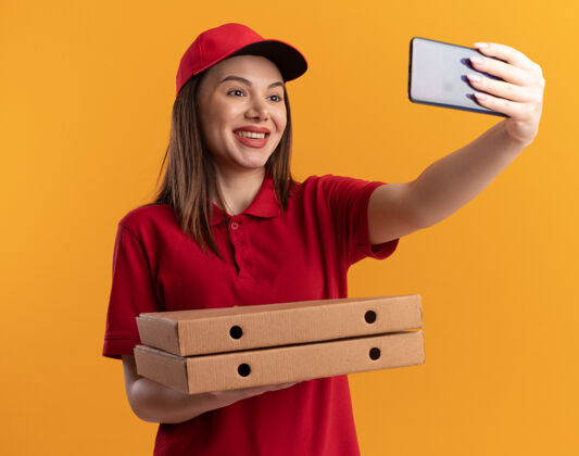 拿着身着制服的微笑漂亮的送货员拿着比萨饼盒 看着手机自拍盒子送货披萨