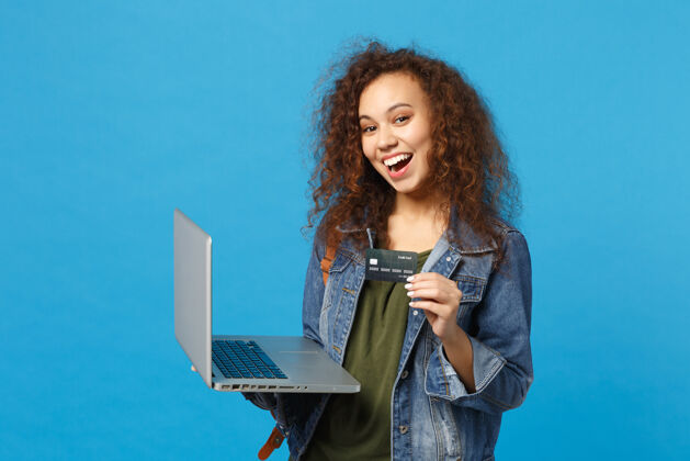 包年轻的非洲裔美国女孩十几岁的学生在牛仔服装 背包工作的电脑 持有银行卡隔离在蓝色的墙壁上支付获奖者青少年