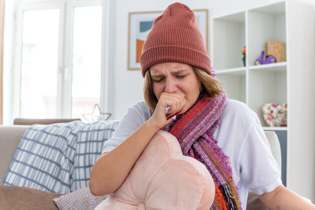痛苦不健康的年轻妇女戴着暖和的帽子 戴着围巾 看起来不舒服 生病 拿着纸巾 患感冒和流感 坐在客厅的沙发上咳嗽围巾咳嗽不安