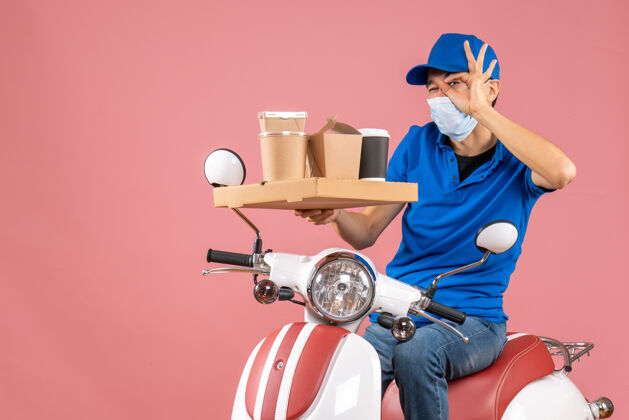 人物男性送货员戴着口罩 戴着帽子 坐在滑板车上送货 在柔和的桃色背景下做眼镜手势交付背景男性