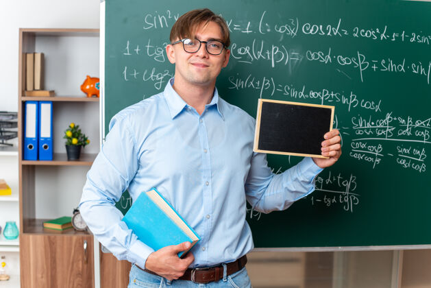站年轻的男老师戴着眼镜拿着书拿着小黑板微笑着自信地站在教室里数学公式的黑板旁教室黑板穿