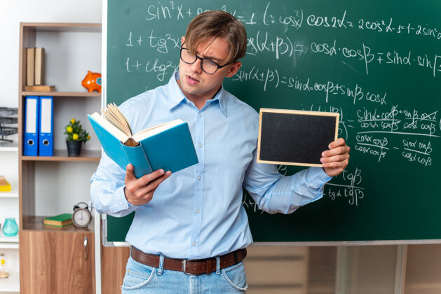 公式年轻的男老师戴着眼镜拿着小黑板看书 自信地站在教室里的数学公式黑板旁教室阅读站
