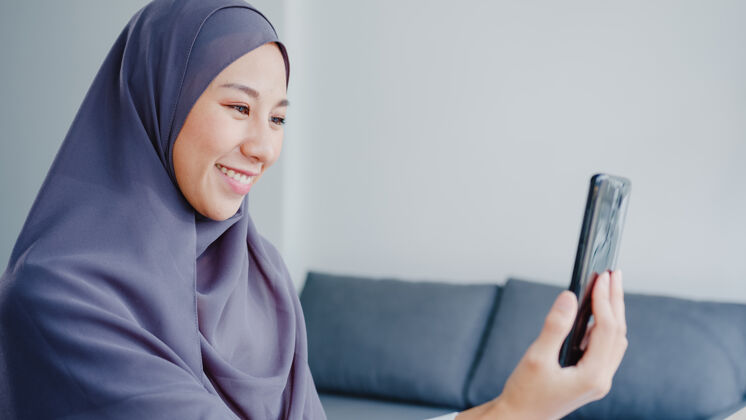 住宅年轻的亚洲女商人使用智能手机通过视频聊天与朋友交谈头脑风暴在线会议 同时在客厅远程工作会议视频通话沙发