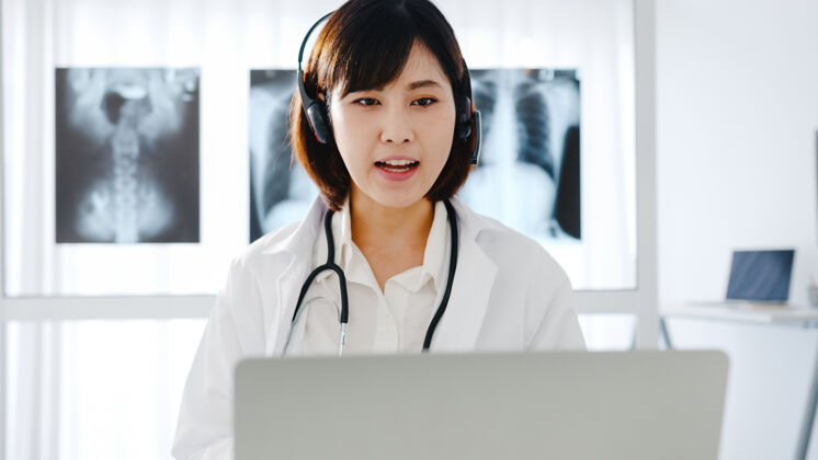 住宅年轻的亚洲女医生 身穿白色医疗制服 手持听诊器 使用笔记本电脑 在诊所或医院的办公桌上与患者进行视频会议呼叫医生咨询