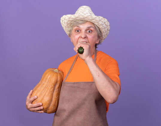 女饥饿的上了年纪的女园丁戴着园艺帽 手里拿着南瓜 咬着黄瓜老人园艺饿
