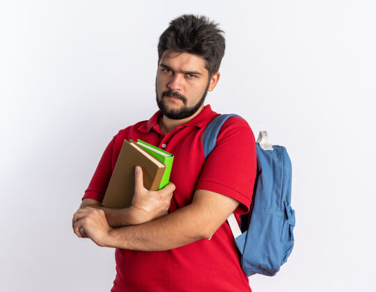 年轻人年轻的留着胡子的学生 穿着红色马球衫 背着背包 手里拿着笔记本 皱着眉头站在白墙上手势背包姿势