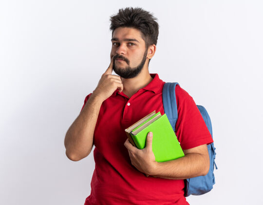 站立年轻的留着胡子的学生 穿着红色马球衫 背着背包 拿着笔记本 站在白色的墙上年轻人马球胡须