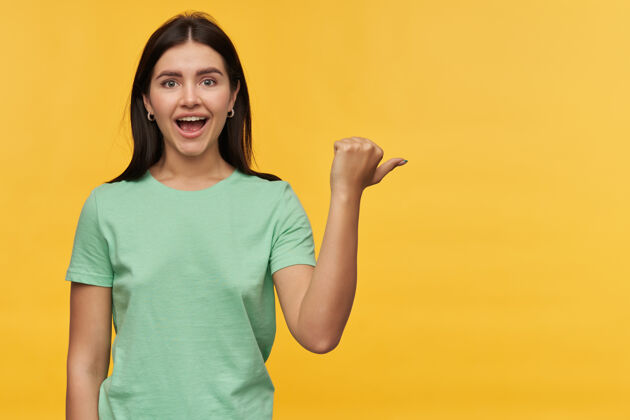 拇指穿着薄荷色T恤 一头黑发的年轻女子站着 用大拇指指着广告空间的一边 隔着黄色的墙壁奇迹成人指指点点