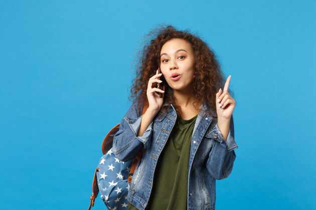 青少年年轻的非洲裔美国女孩十几岁的学生在牛仔服装 背包举行电话隔离在蓝色的墙壁上设备牛仔裤电话