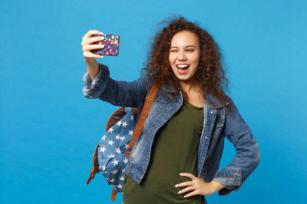人年轻的非洲裔美国女孩十几岁的学生在牛仔服装 背包举行电话隔离在蓝色的墙壁上手机站立姿势