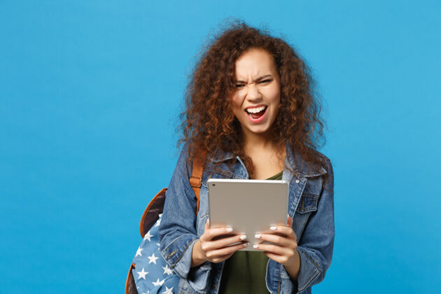 女人年轻的非洲裔美国女孩十几岁的学生在牛仔服装 背包举行垫电脑隔离在蓝色的墙壁上学生青少年人