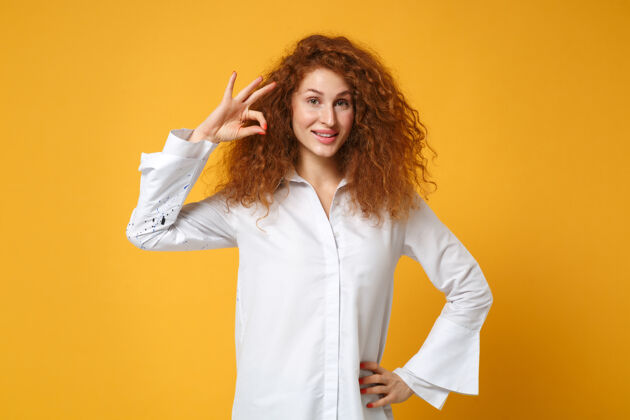 手指微笑着的年轻红发女子穿着休闲白衬衫在黄橙色的墙上摆出孤立的姿势卷发肖像女性