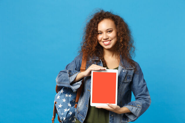 学生年轻的非洲裔美国女孩十几岁的学生在牛仔服装 背包举行垫电脑隔离在蓝色的墙壁上青少年显示器复制空间