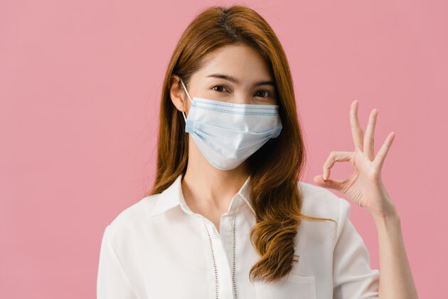 过敏年轻的亚洲女孩戴着医用面罩 穿着休闲服 打着ok的手势 看着隔离在粉色背景上的相机卫生学生医学