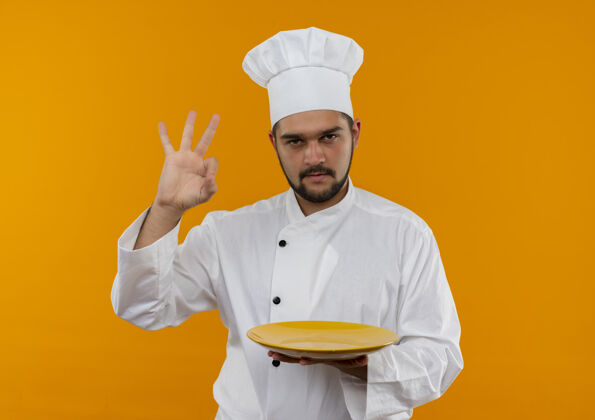 好的自信的年轻男性厨师 身着厨师制服 手拿空盘子 在橙色的墙上贴着“做得好”的牌子 上面留有复印空间盘子年轻烹饪