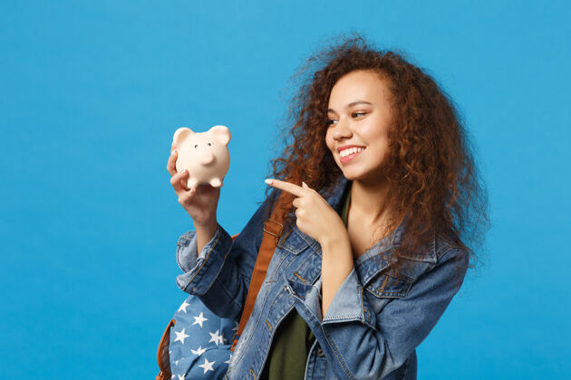 付款年轻的非洲裔美国女孩十几岁的学生在牛仔服装 背包举行猪隔离在蓝色的墙壁上粉红色奖经济