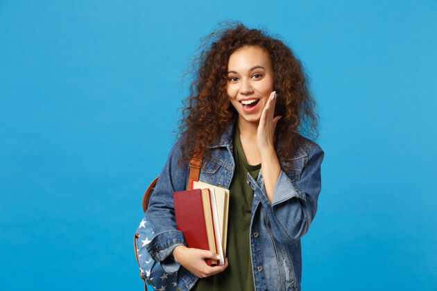 知识穿着牛仔服 背着书包的年轻女学生把书孤立地放在蓝色的墙上女复制空间班级