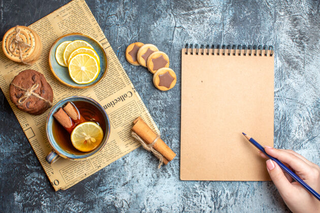 笔上图：美味的饼干和一杯红茶加肉桂放在旧报纸上 拿着钢笔放在黑色背景的螺旋笔记本上茶笔记本美味的饼干