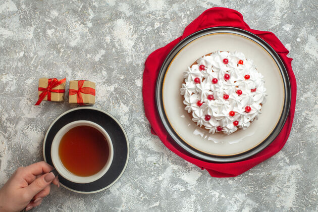过滤器上图：美味的奶油蛋糕上点缀着水果 红色毛巾上手拿一杯红茶 小礼盒上放着冰背景水果杯子毛巾
