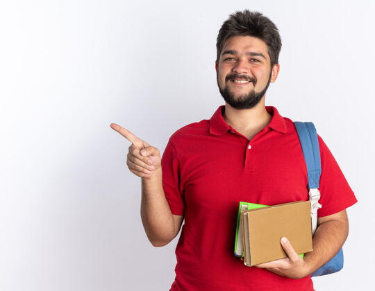 指向快乐的年轻留着胡子的学生 穿着红色马球衫 背着背包 拿着笔记本 愉快地微笑着 用食指指着站在白墙上的一边马球手指笔记本