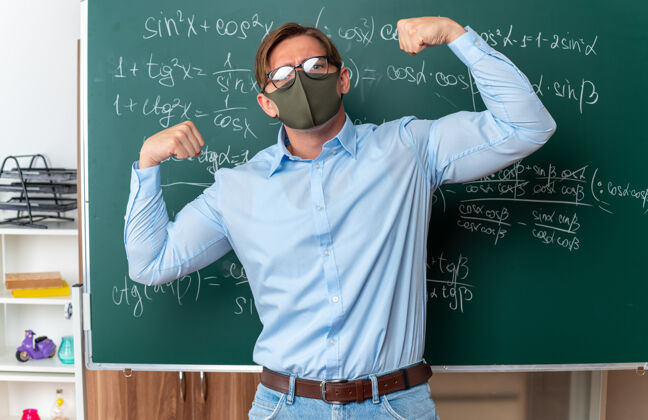 教室戴着防护面具戴着眼镜的年轻男老师举起拳头 像一个胜利者一样自信地站在教室里的数学公式黑板旁戴数学拳头