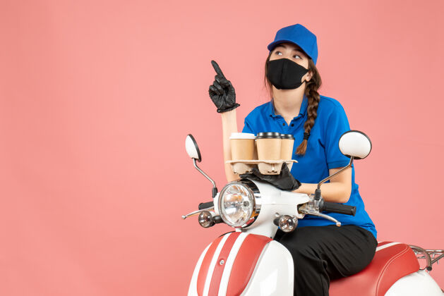 女性前视图好奇的女性送货员戴着医用口罩和手套坐在滑板车上拿着订单指着粉彩桃背景桃奇迹医疗