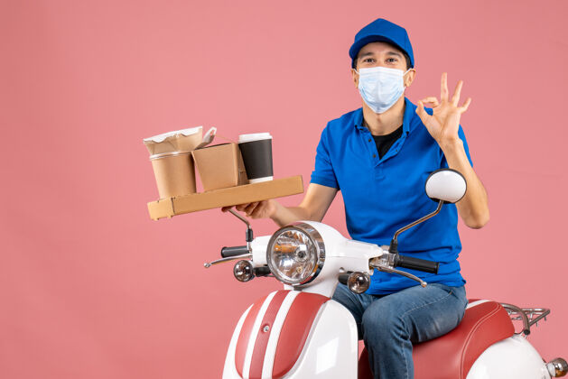 帽子俯视图微笑的快递员戴着医用面罩戴着帽子坐在滑板车上做着戴眼镜的手势在粉彩桃上人面具医学