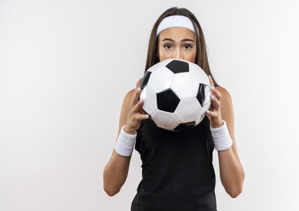 印象令人印象深刻的漂亮运动女孩戴着头带和腕带拿着足球 躲在它后面的白色墙壁上与复制空间隔离头带球隐藏