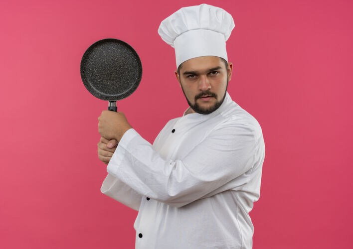 厨师自信的年轻男厨师身着厨师制服 手拿煎锅孤立地站在粉红色的墙上年轻烹饪持有