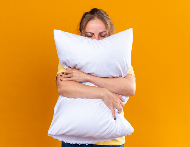 感觉不健康的女人穿着黄色衬衫 患流感和感冒 感觉不舒服 抱着枕头站在橙色的墙上睡觉感冒流感枕头