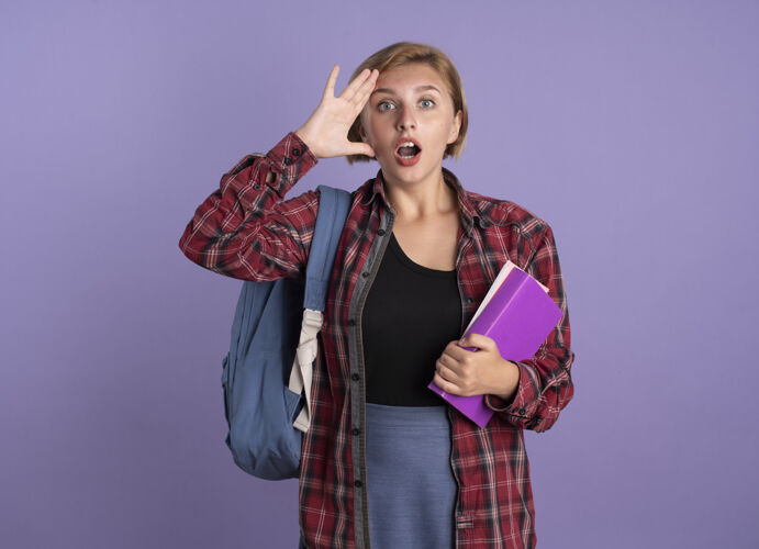 学生惊讶的年轻斯拉夫学生女孩背着背包把手放在额头上拿着书和笔记本举行空间紫色