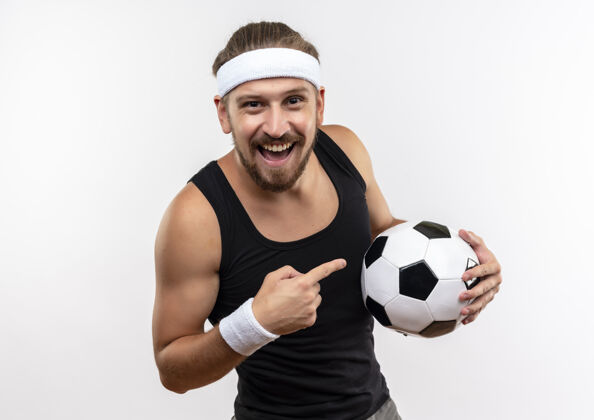 年轻快乐的年轻英俊的运动型男子戴着头带和腕带拿着足球指着隔离在白墙上头带持有足球