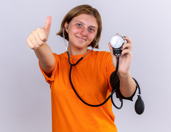 压力不健康的年轻女子穿着橙色t恤 用眼压计测量血压 脸上带着微笑 站在白墙上竖起大拇指显示使用脸