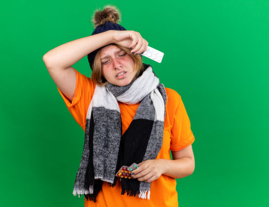 围巾不健康的年轻女子穿着橙色t恤 戴着帽子 脖子上围着暖和的围巾 感觉很可怕 拿着药片 忍受着病毒 发烧站在绿色的墙上抱可怕不健康