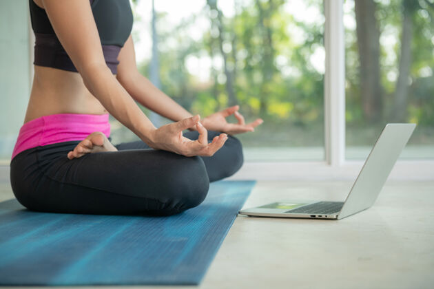 活跃运动型女人练习瑜伽 做阿达帕德玛萨纳运动 莲花式冥想 锻炼 穿着运动服 在笔记本电脑上看在线健身视频教程 坐在家里锻炼锻炼课程房间