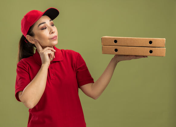 披萨身穿红色制服 头戴鸭舌帽 手持比萨盒的年轻送货员站在绿色的墙上 带着怀疑的表情看着比萨盒年轻站着盒子
