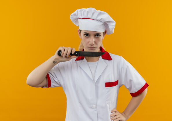 拿着自信的年轻漂亮厨师 身着厨师制服 手放在腰间拿着刀 隔离在橙色的墙上厨师刀手