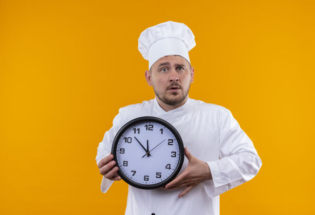印象深刻令人印象深刻的年轻帅哥厨师在厨师制服举行的橙色墙上孤立的时钟制服手持年轻
