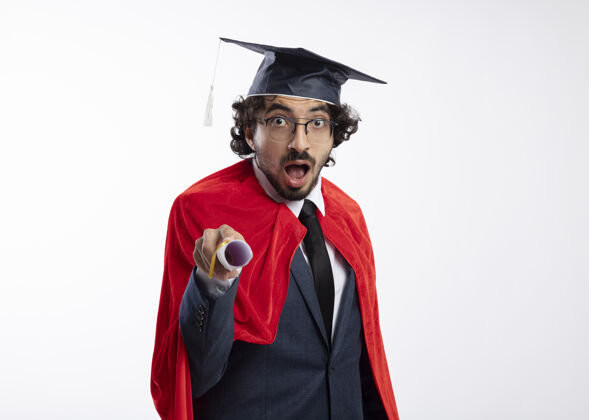 毕业惊讶的年轻白种人超级英雄戴着眼镜 穿着红色斗篷的西装 戴着毕业帽 手里拿着毕业证书证书西装持有