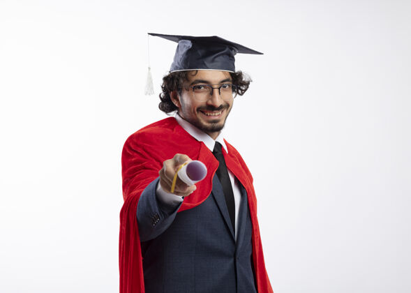 太空年轻的高加索超级英雄面带微笑 戴着眼镜 身穿红色斗篷的西装 戴着毕业帽 手里拿着毕业证书斗篷红色西装
