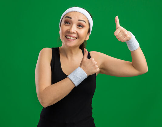 健康戴着头带和臂章 脸上带着微笑 竖起大拇指 站在绿色墙壁上的快乐的年轻健身女士站臂章头带