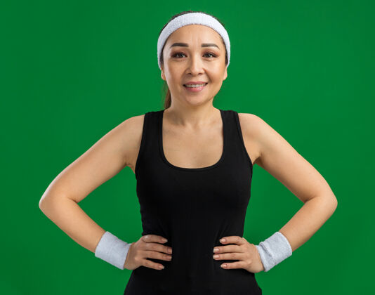 站立戴着头带和臂章的快乐的年轻健身女士微笑着自信地站在绿色的墙上自信臂章头带