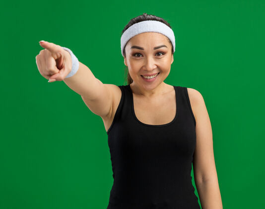 目录戴着头带和臂章的年轻健身女士微笑着自信地用食指指着绿色的墙壁女人指点健身