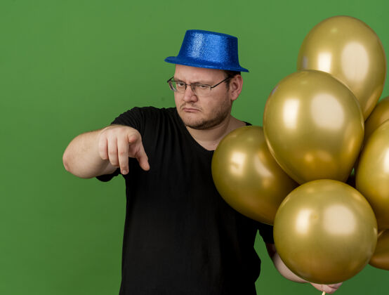 聚会自信的成年斯拉夫男子戴着眼镜 头戴蓝色派对帽 手持氦气球 指着摄像机穿点举行